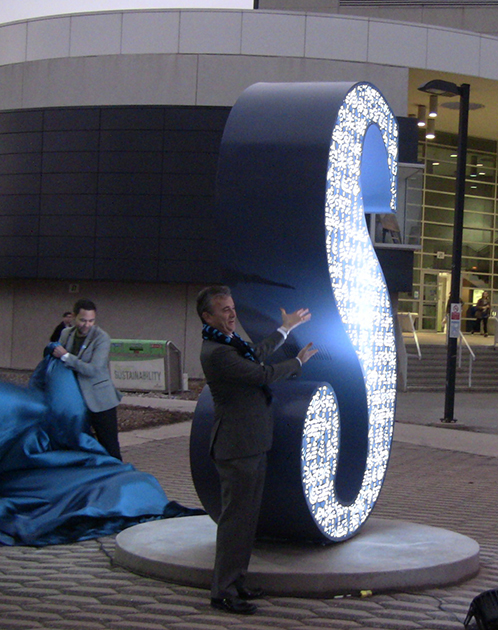 President Jeff Zabudsky standing next to the glowing ‘S’ sculpture moments after its artist, Pierre Poussin, removes the veil. 